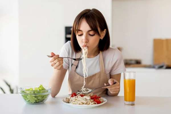 Young housewife tasting spaghetti and vegetable salad, enjoying delicious lunch at home, sitting in modern kitchen — Stock Photo, Image