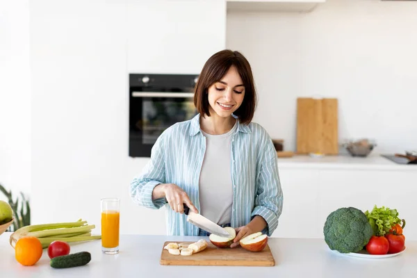 Fröhliche junge Frau schneidet frische Äpfel auf Holzbrett, während sie Obstsalat zum Frühstück in der Küche zubereitet — Stockfoto