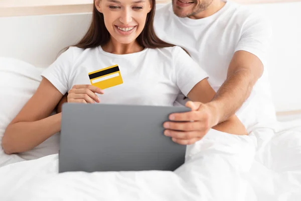 Vágott mosolygós kaukázusi fiatal család ölelés, ül az ágyban, és a hitelkártya és laptop vásárlás — Stock Fotó