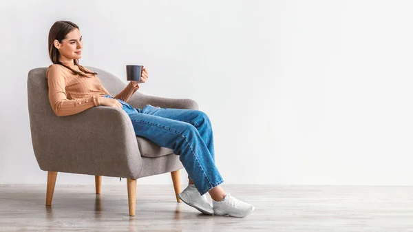 Full längd av tusenårig kvinna dricker färskt kaffe, har paus i bekväma fåtölj mot vit vägg, panorama — Stockfoto
