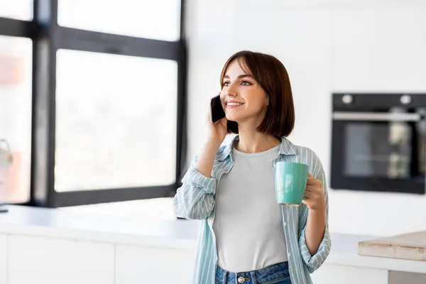 Jonge mooie dame praten op mobiele telefoon en het drinken van koffie in de ochtend terwijl staan in licht moderne keuken interieur — Stockfoto
