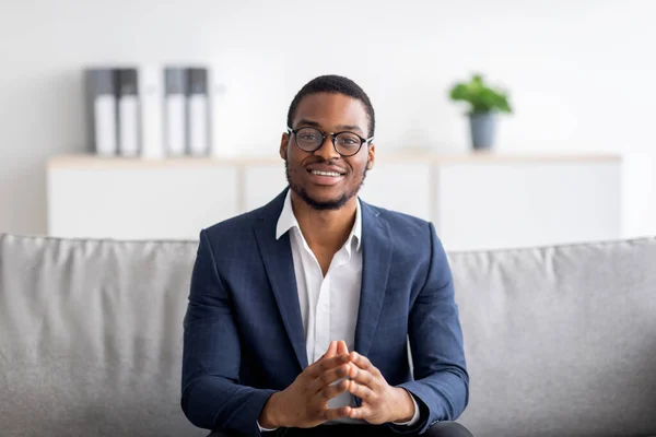 Porträt eines fröhlichen jungen schwarzen Psychologen, der in die Kamera lächelt und auf dem Sofa in der Klinik sitzt. Konzept für psychische Gesundheit — Stockfoto