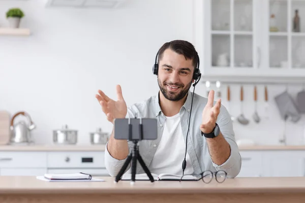 Sonriente adulto europeo hombre profesor en los auriculares gestos y se ve en el teléfono inteligente webcam en el interior de la cocina — Foto de Stock