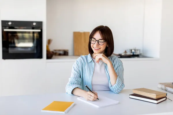 Lleva un diario, escribe pensamientos e ideas. Jovencita soñadora sentada en cocina y escribiendo en cuaderno con bolígrafo —  Fotos de Stock