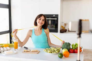 Mutlu kadın fitness vlog kaydedicisi akıllı telefondan video blogu, yeme alışkanlıkları hakkında tavsiye paylaşımı, mezura ile ayakta durmak