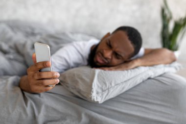 Kafası karışmış siyah adam yatakta telefona bakıyor.