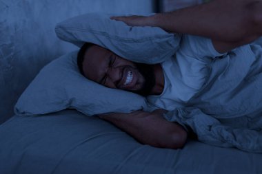 Stressed black man covering ears lying in bed at hight clipart