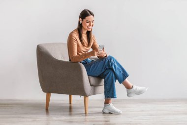 Kulaklıklı mutlu beyaz kadın akıllı telefon kullanıyor, online sohbet ediyor, koltukta oturuyor, sosyal medyada paylaşıyor.