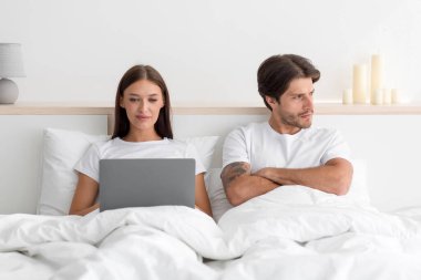 Kırılmış genç Avrupalı koca kollarını kavuşturup karısına sırt çevirmiş, dizüstü bilgisayarında sohbet eden bir kadın.