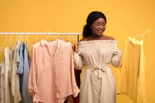 Mulher negra em pé perto de rack de roupas com roupas elegantes, escolhendo entre duas roupas, compras para guarda-roupa na moda — Fotografia de Stock