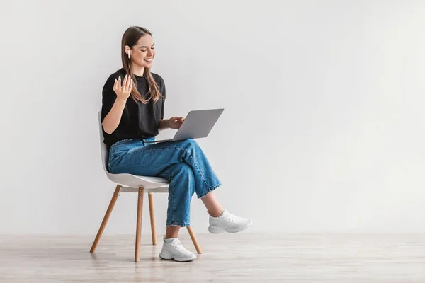 Cheerful young woman in earphones using laptop, having online meeting, sitting on chair against white wall, empty space