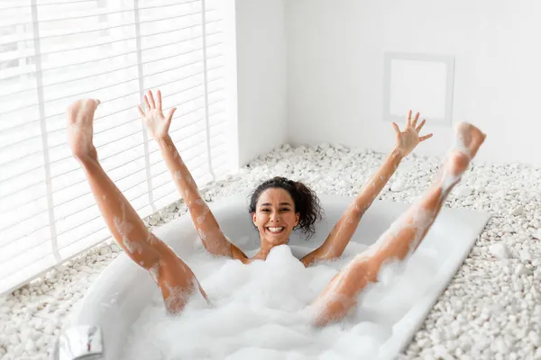 Jovem tola se divertindo no banho de bolhas, levantando os braços e pernas, sendo boba e brincalhona em casa — Fotografia de Stock