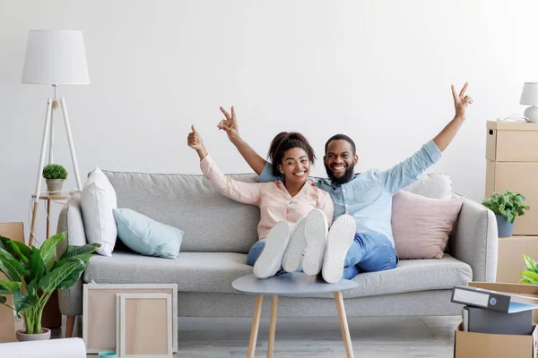 Sorrindo animado jovem casal negro comemorar dia em movimento em sua própria casa, senta-se no sofá, mostra sinal de paz e polegar para cima — Fotografia de Stock