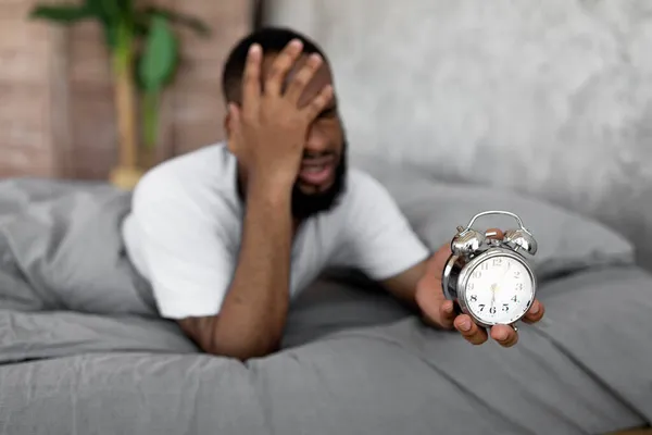 Hombre negro sufriendo de insomnio sosteniendo reloj en la cama — Foto de Stock