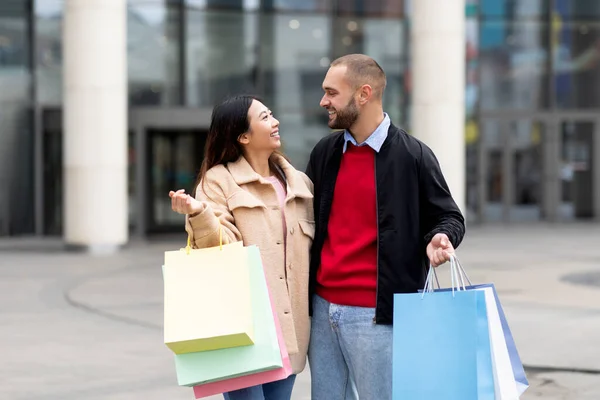 Millennial multiraciale paar staan samen in de buurt van winkelcentrum, het dragen van boodschappentassen, kijken naar elkaar met genegenheid — Stockfoto