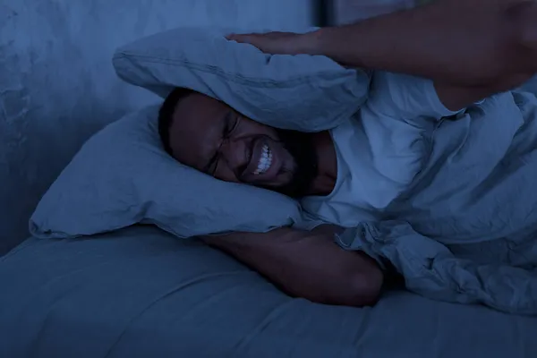 Stressed black man covering ears lying in bed at hight