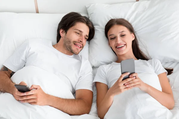 Alegre guapo millennial europeo macho mira esposa smartphone en la cama en el interior del dormitorio — Foto de Stock