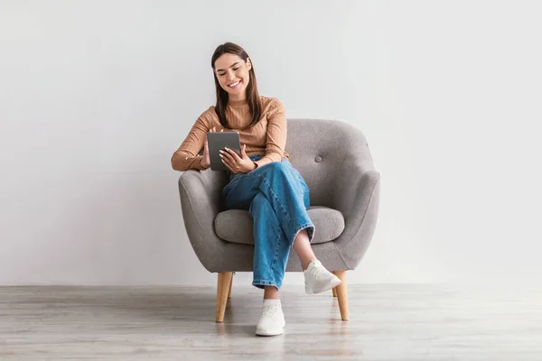 Θετική νεαρή γυναίκα που εργάζεται online, κάθεται σε πολυθρόνα με ψηφιακή ταμπλέτα ενάντια σε λευκό τοίχο στούντιο, πλήρους μήκους — Φωτογραφία Αρχείου
