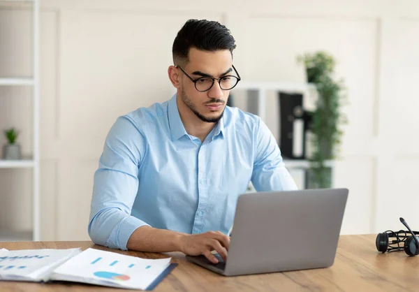 Seriöser junger Araber, der am Schreibtisch mit Laptop arbeitet und im Büro auf der Tastatur tippt — Stockfoto