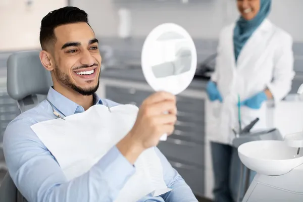 Homem árabe bonito feliz que olha para o espelho após o tratamento dos dentes na clínica — Fotografia de Stock