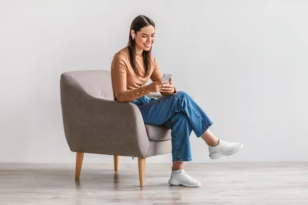 Mujer caucásica feliz con auriculares usando el teléfono inteligente, tener chat en línea, sentarse en el sillón, publicar en las redes sociales —  Fotos de Stock