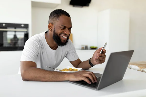 Black man using pc and smartphone having breakfast at home — Stock Photo, Image