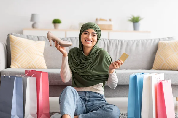Elinde kredi kartı ve yeni ayakkabıyla tesettürlü Arap bir kadın hediye paketlerinin arasında yerde oturuyor, evde internetten ayakkabı alıyor. — Stok fotoğraf