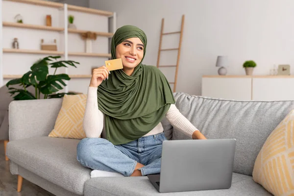 Uzaktan ödeme için kredi kartı kullanan tesettürlü bir Arap kadın, dizüstü bilgisayarla alışveriş yapan, evdeki web mağazasından mal alan bir kadın. — Stok fotoğraf