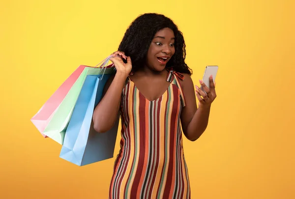 Scioccato signora nera con borse della spesa guardando smartphone, utilizzando l'applicazione mobile, l'acquisto di cose online — Foto Stock