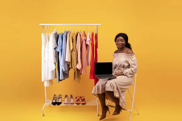 Linda mulher negra sentada perto do rack de roupas, segurando laptop com tela vazia, oferecendo maquete para design de loja web — Fotografia de Stock
