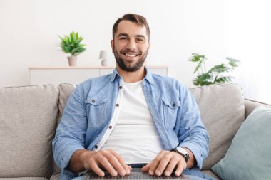 Keyifli, orta yaşlı, rahat bir şekilde kanepede dinlenen beyaz adam laptopunda video görüşmesi yapıyor.