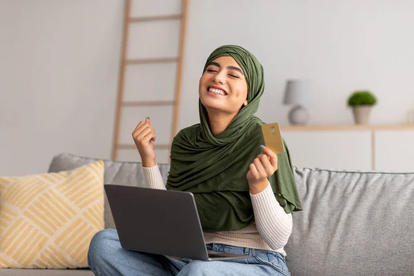 Giovane donna araba emotiva in hijab in possesso di carta di credito, utilizzando il computer portatile, facendo gesto SI, shopping online a casa — Foto Stock