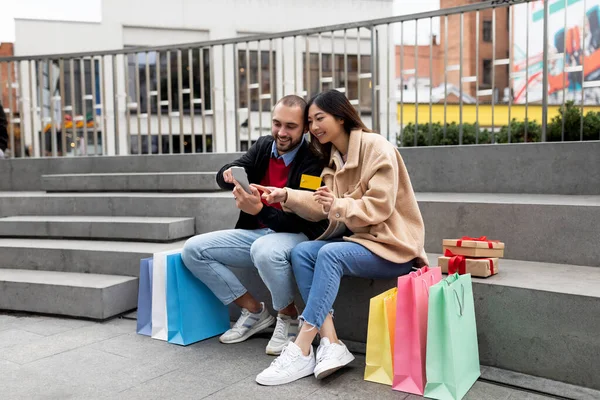 Merdivenlerde oturan genç çift akıllı telefon ve kredi kartıyla, internetten alışveriş yapıyorlar, alışveriş torbaları ve hediyelerle çevrili. — Stok fotoğraf