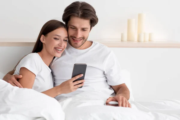 Feliz joven pareja caucásica ver vídeo o chatear en las redes sociales en el teléfono inteligente, abrazos en la cama — Foto de Stock