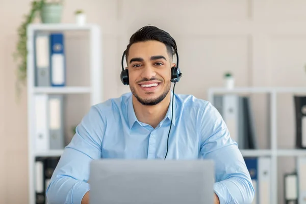 Communication à distance. Homme arabe joyeux avec casque de travail en ligne sur ordinateur portable, ayant appel vidéo au bureau — Photo