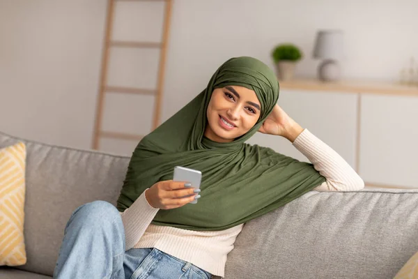 Lächelnde Araberin im Hijab sitzt auf dem Sofa, nutzt Smartphone, surft im Internet, chattet online, schaut sich zu Hause Filme an — Stockfoto