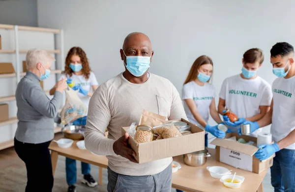 Charity organization. African american senior man holding box with donations food, wearing medical mask