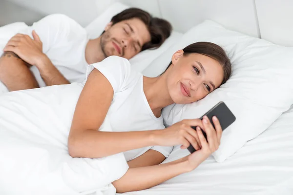 Joven marido caucásico durmiendo, esposa sonriente mirando el teléfono, charlando con el amante, navegar en Internet en la cama — Foto de Stock