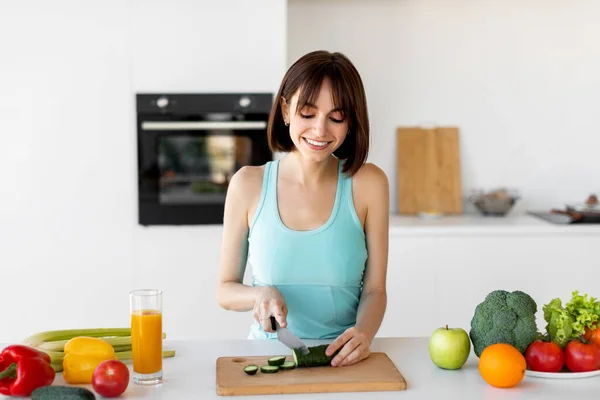 Detox recipes. Slim woman cooking healthy vegetable salad, cutting cucumber on wooden board, standing in kitchen