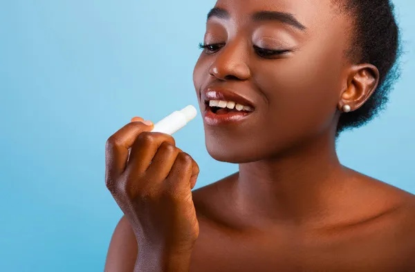 Bibir peduli. Wanita muda african Amerika menerapkan balsem bibir, pelembab merawat kulit, latar belakang biru, closeup — Stok Foto