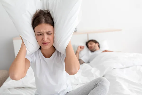 Unhappy offended angry young caucasian female sitting on bed, covering her ears with pillow — Stock Photo, Image