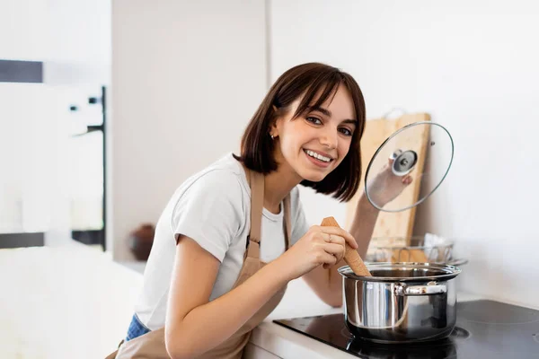 Glückliche Dame, die Suppe probiert, während sie in einem Topf Abendessen kocht, in der modernen Küche steht und mit Schürze in die Kamera lächelt — Stockfoto