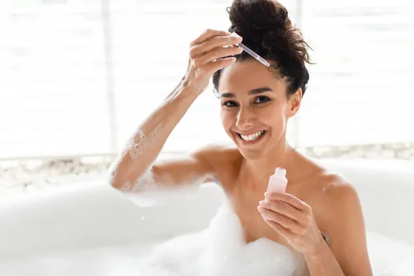 Jovem aplicando soro hidratante no rosto enquanto relaxa no banho espumoso, fazendo tratamento de beleza em casa — Fotografia de Stock