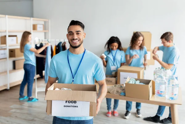 Mladý arabský dobrovolník drží kartónovou krabici na dárcovství a usmívá se před kamerou, pracuje v charitativním centru — Stock fotografie