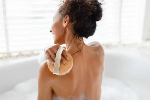 Back view of young woman making lymphatic massage with brush, scrubbing her skin in foamy bath at home