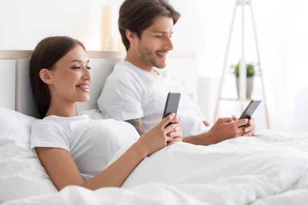 Vrolijke jonge blanke man en vrouw kijken video of chatten in sociale netwerken in smartphones in bed — Stockfoto