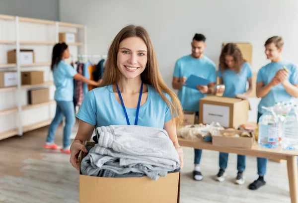 Happy female volunteer holding box with clothes for donation, smile to camera while working at charity center — Stock fotografie