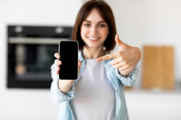 Felice signora mostrando schermo smartphone vuoto e puntando su di esso mentre in piedi in cucina interno, messa a fuoco selettiva — Foto Stock