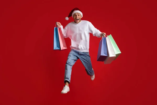 Vánoční prodej. Vtipný vzrušený muž v Santa klobouku skákání s nákupními taškami — Stock fotografie
