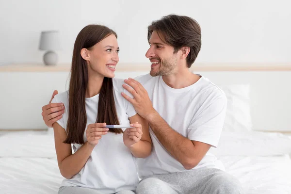 Lächelndes europäisches Ehepaar freut sich über Ergebnis des Schwangerschaftstests, sitzt im Bett — Stockfoto